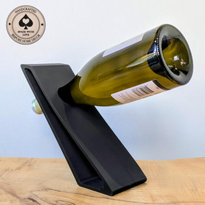 A black 3d printed wine bottle holder on a light wood surface against a pale blue background, holding a green glass wine bottle. A circular badge in the top-left reads 'Handcrafted Spade with Love' and 'Bespoke Home Decor'.