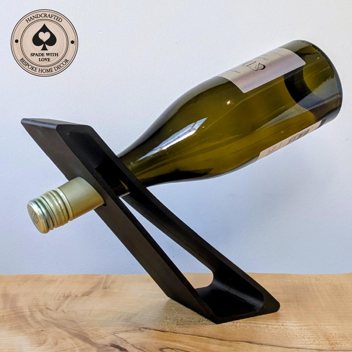 A black 3d printed wine bottle holder on a light wood surface against a pale blue background, holding a green glass wine bottle. A circular badge in the top-left reads 'Handcrafted Spade with Love' and 'Bespoke Home Decor'.