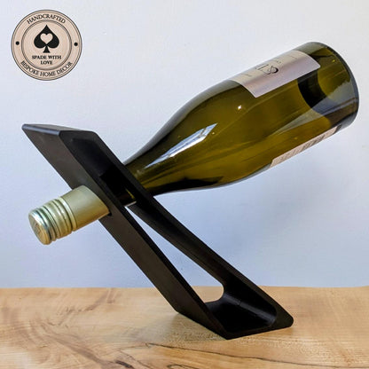 A black 3d printed wine bottle holder on a light wood surface against a pale blue background, holding a green glass wine bottle. A circular badge in the top-left reads 'Handcrafted Spade with Love' and 'Bespoke Home Decor'.