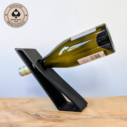 A black 3d printed wine bottle holder on a light wood surface against a pale blue background, holding a green glass wine bottle. A circular badge in the top-left reads 'Handcrafted Spade with Love' and 'Bespoke Home Decor'.